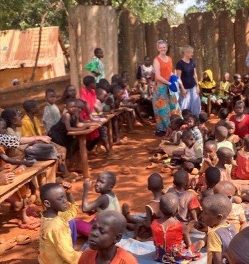 Kinder sitzen auf dem Boden und an Holzbänken und warten auf das Essen.
