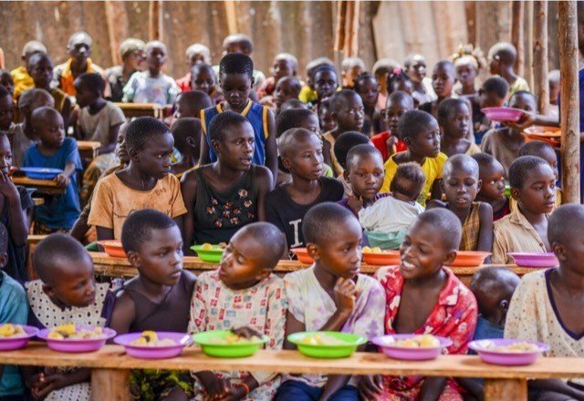 Viele Kinder an langen Bänken, vor ihnen bunte Teller mit Essen.