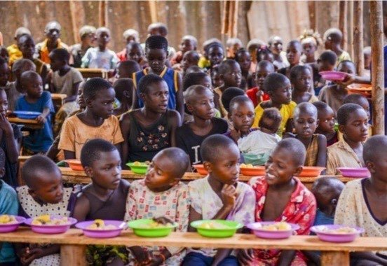 Viele Kinder an langen Bänken, vor ihnen bunte Teller mit Essen.